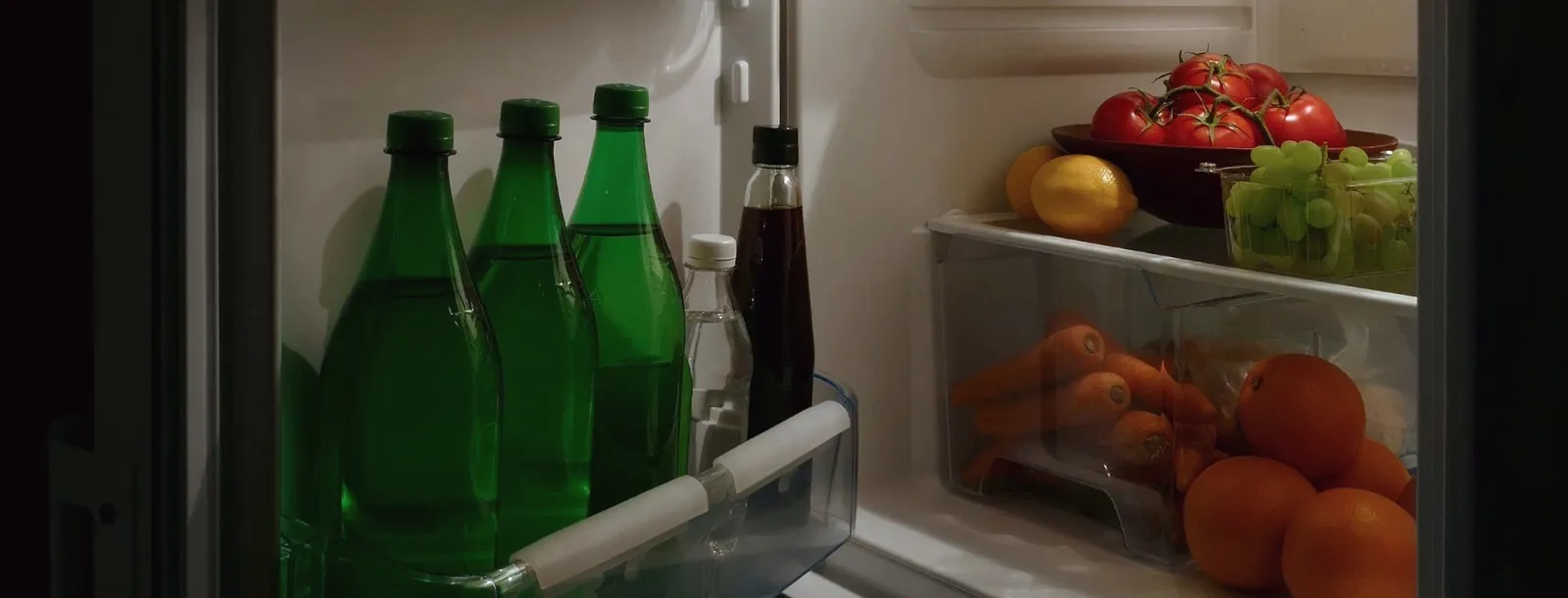 An open fridge at night, shelves holding tomatoes, grapes, carrots and oranges.